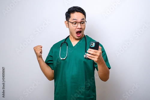 Shocked asian male doctor, nurse in scrubs and stethoscope screaming proud and celebrating victory while holding smart phone