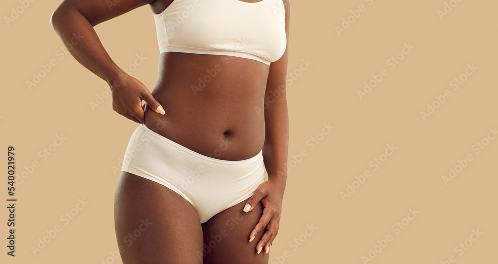 Black girl in underwear pinches belly side fat standing isolated on beige color background. Plus size woman fights excessive body weight with exercising, eating less or doing laser lipolysis procedure - obrazy, fototapety, plakaty 