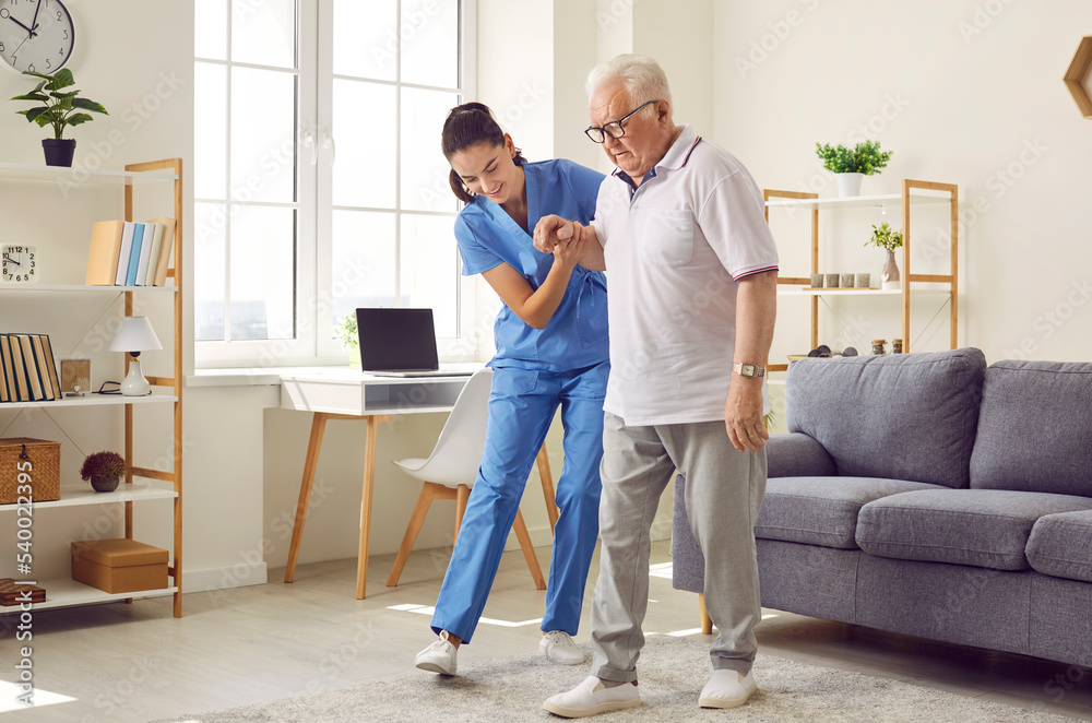 © Studio Romantic - Young nurse helping elderly man walk in the room, holding his hand, supporting him. Treatment and rehabilitation after injury or stroke, life in assisted living facility, senior care concept