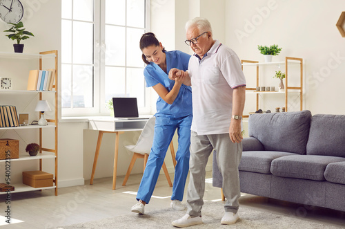 Young nurse helping elderly man walk in the room, holding his hand, supporting him. Treatment and rehabilitation after injury or stroke, life in assisted living facility, senior care concept