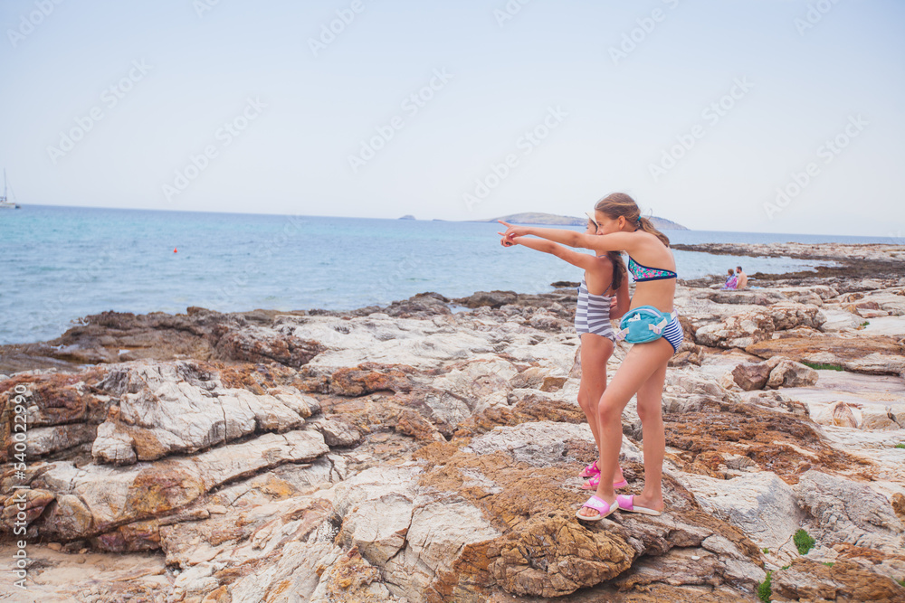 Two little girls on rocky shore watch beautiful seascape on summer day at vacation.