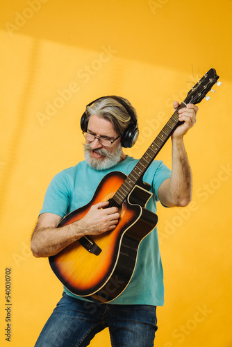 Senior man in glasses and headphones playing the guitar on a yellow background. Studio shooting.