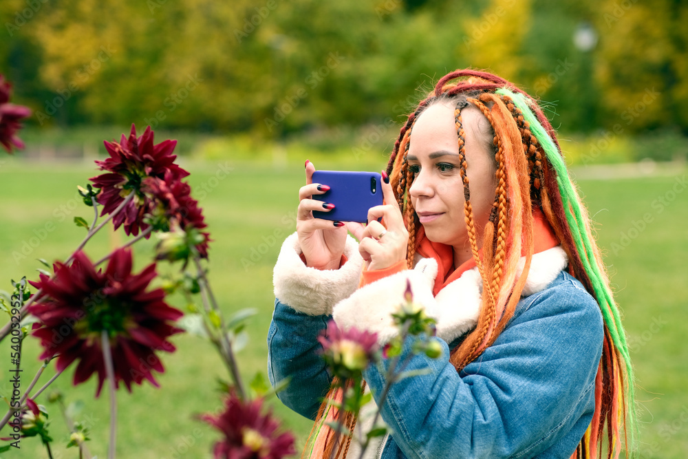 Woman taking pictures on smartphone in park. Positive young female hipster with long dreadlocks in casual clothes photographing flowers on mobile phone in park Woman taking pictures on smartphone in park. Positive young female hipster with long dreadlocks in casual clothes photographing flowers on mobile phone in park