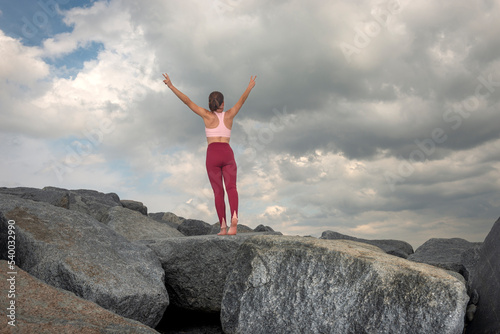 Sporty woman stands on top of rocks after a climb, arms raised in celebration.