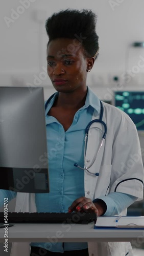 FVertical video: ront view of afro american doctor typing medical expertise on computer while in background nurse checking heartbeat pulse using oximeter. Hospitalized patient having respiratory photo