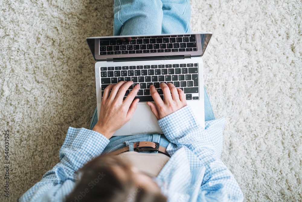Fototapeta premium Crop photo of young woman in jeans working on laptop sitting on carpet at home, view from top