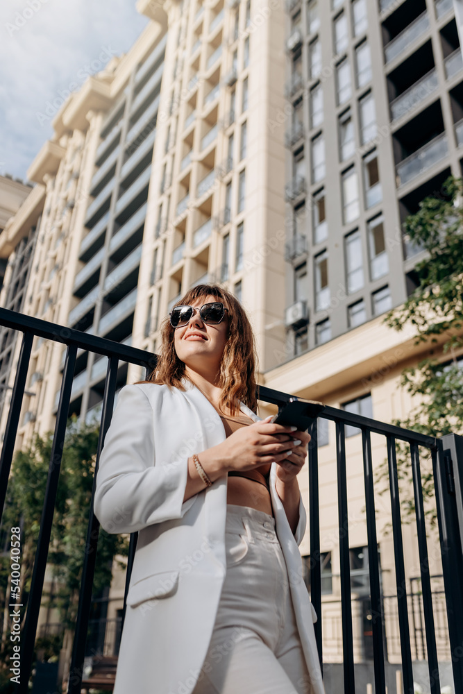 Smiling businesswoman in white suit using phone during walking in city with modern architecture and looking at side