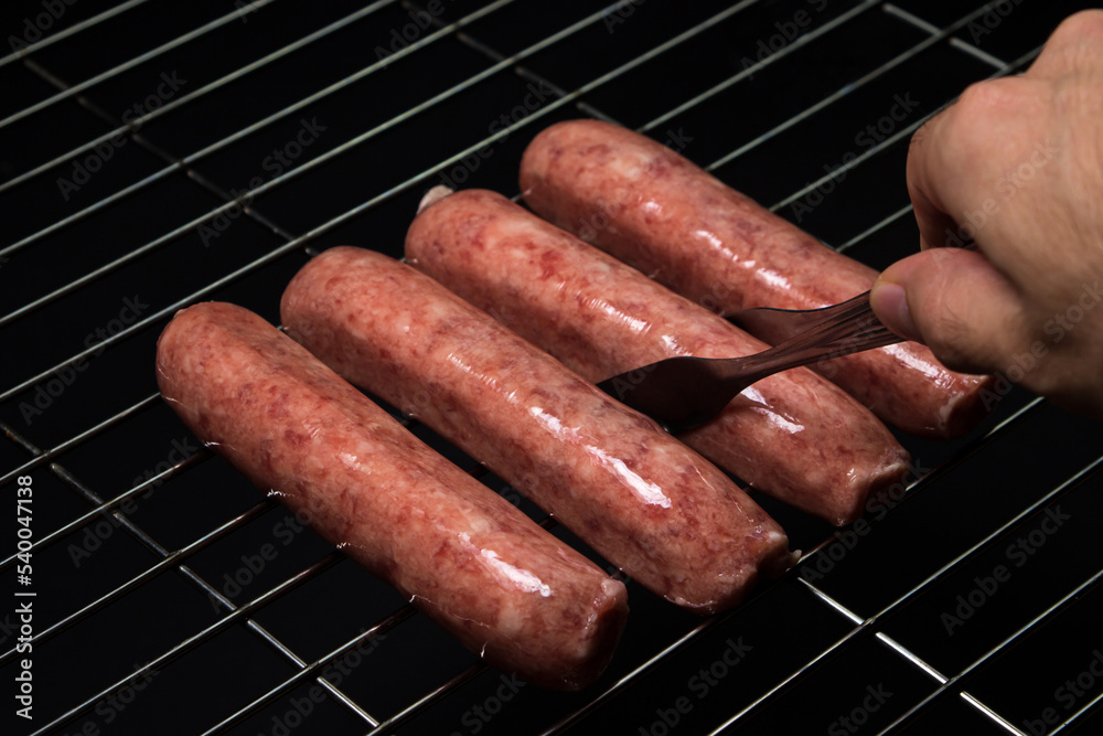 Raw sausages on a grill grate on a black background. Meat products at home. Tongs take sausage on the grill Raw sausages on a grill grate on a black background. Meat products at home. Tongs take sausage on the grill