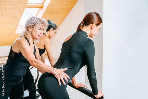 Elderly female instructor helping young woman in doing pilates workout at studio.
