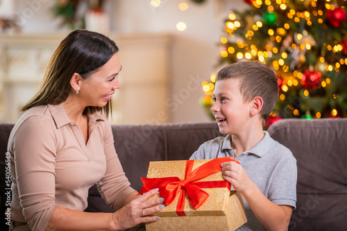 happy loving mother and son hugging at home, sitting on sofa against background Christmas tree. child and woman laughing happily, holding gift box in their hands. Give each other gifts at Christmas