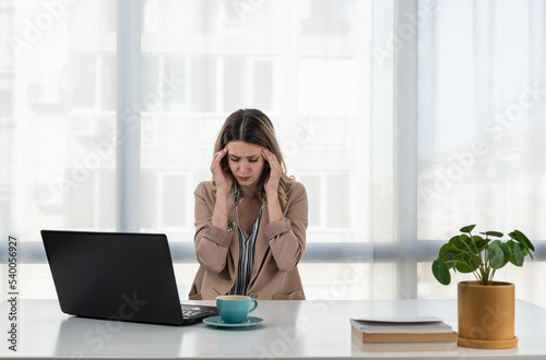 Exhausted upset young business woman sitting at table with laptop, leaning head on hand, suffering from headache, feeling depressed, unhappy, tired, getting bad news. Professional burnout concept