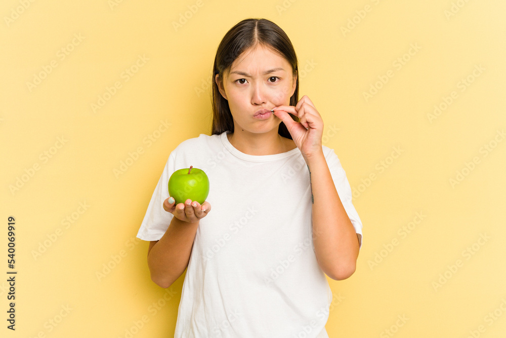 Young Asian woman eating an apple isolated on yellow background with fingers on lips keeping a secret. Young Asian woman eating an apple isolated on yellow background with fingers on lips keeping a secret.