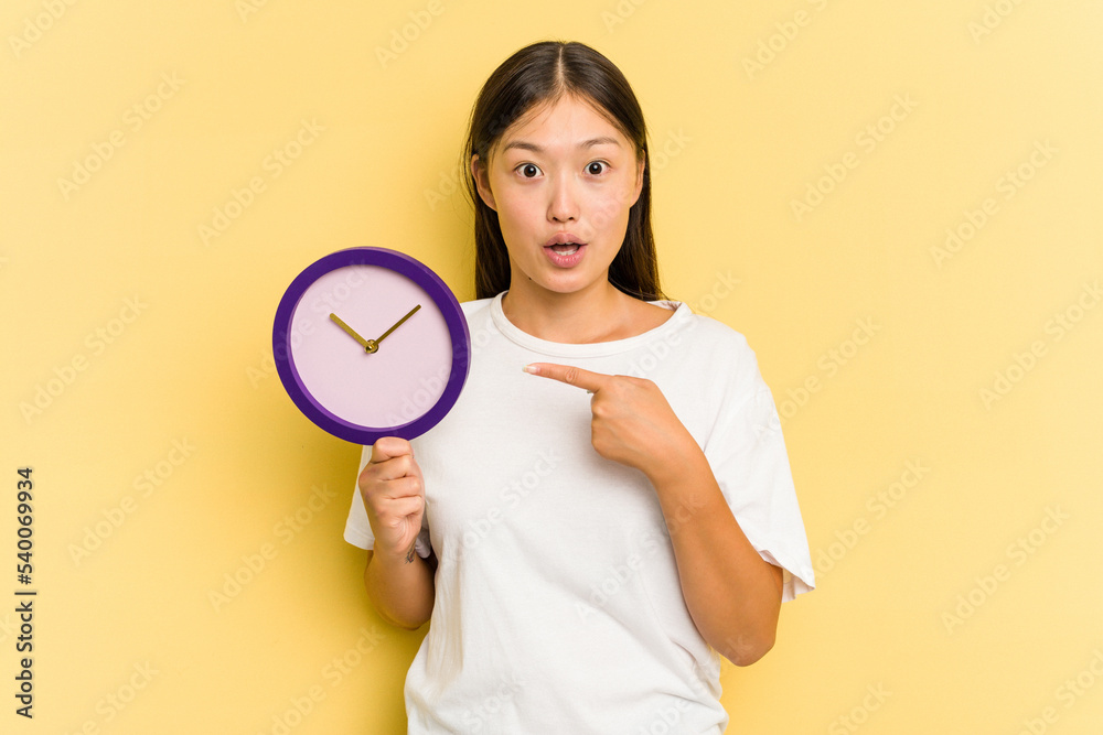 Young asian woman holding a clock isolated on yellow background pointing to the side Young asian woman holding a clock isolated on yellow background pointing to the side