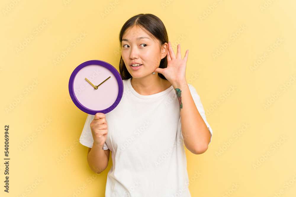 Young asian woman holding a clock isolated on yellow background trying to listening a gossip. Young asian woman holding a clock isolated on yellow background trying to listening a gossip.