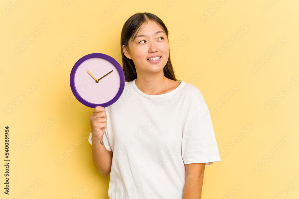Young asian woman holding a clock isolated on yellow background looks aside smiling, cheerful and pleasant. Young asian woman holding a clock isolated on yellow background looks aside smiling, cheerful and pleasant.