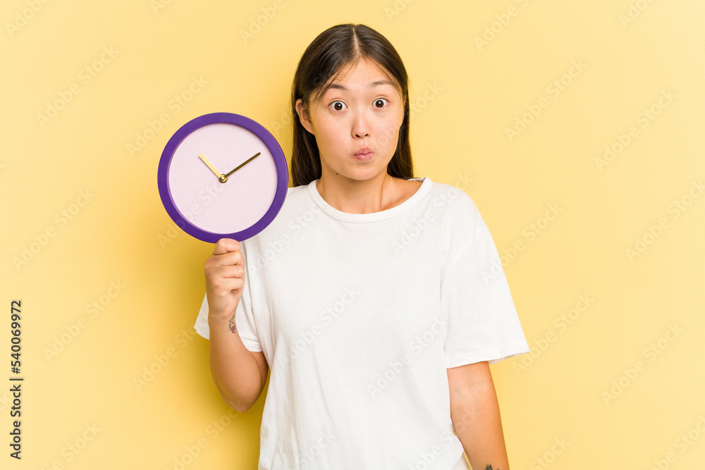 Young asian woman holding a clock isolated on yellow background shrugs shoulders and open eyes confused. Young asian woman holding a clock isolated on yellow background shrugs shoulders and open eyes confused.