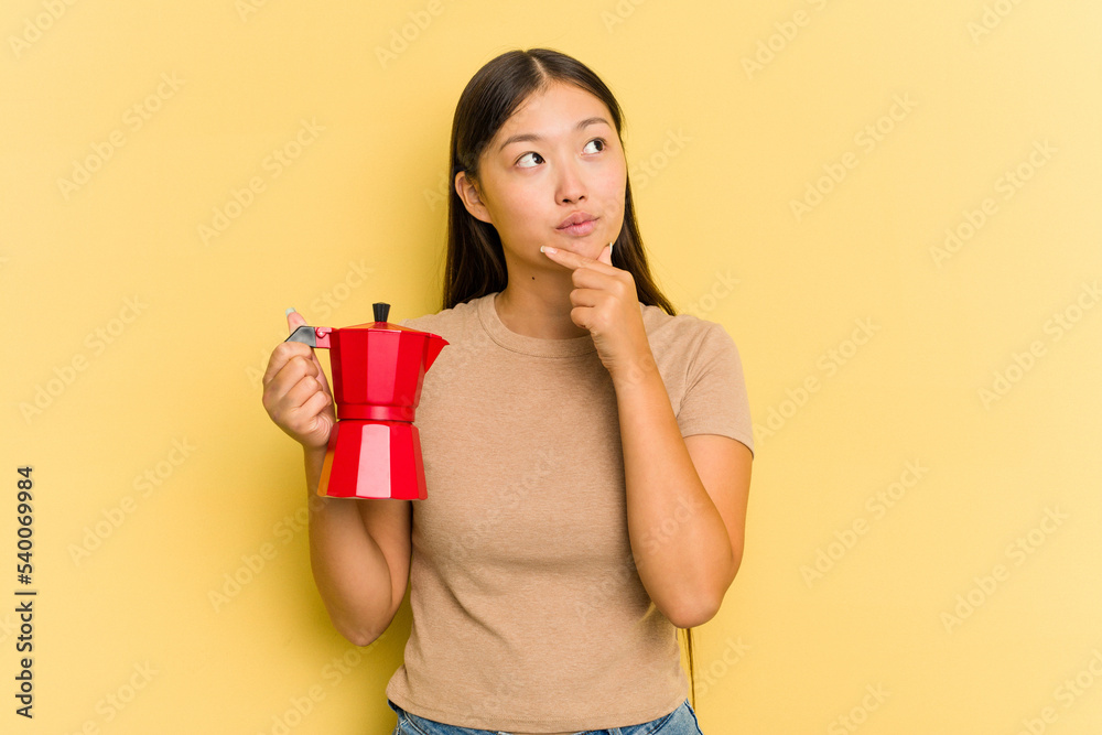 Young Asian woman holding coffee maker isolated on yellow background looking sideways with doubtful and skeptical expression. Young Asian woman holding coffee maker isolated on yellow background looking sideways with doubtful and skeptical expression.