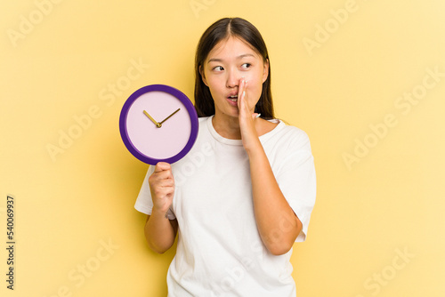 Young asian woman holding a clock isolated on yellow background is saying a secret hot braking news and looking aside