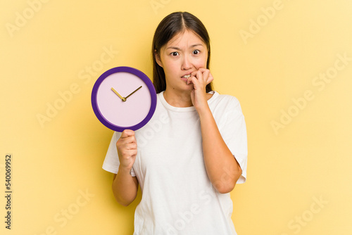 Young asian woman holding a clock isolated on yellow background biting fingernails, nervous and very anxious.