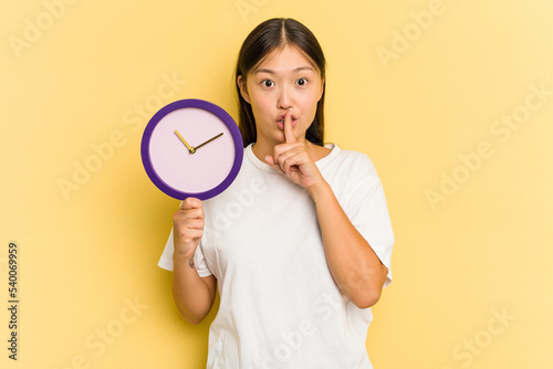 Young asian woman holding a clock isolated on yellow background keeping a secret or asking for silence.