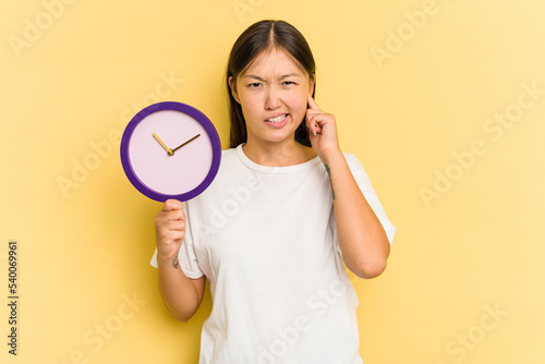 Young asian woman holding a clock isolated on yellow background covering ears with hands.