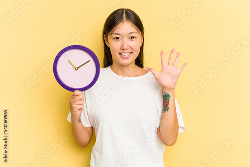 Young asian woman holding a clock isolated on yellow background smiling cheerful showing number five with fingers.