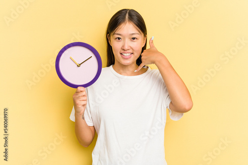 Young asian woman holding a clock isolated on yellow background showing a mobile phone call gesture with fingers.