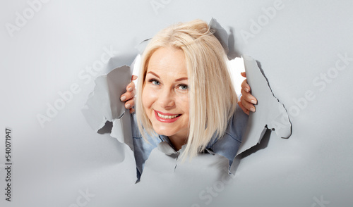 Charismatic joyful woman with blonde hairstyle, looking playfully, wears fashionable hoody, stands in torn paper hole, notices funny pleasant. Gray background.