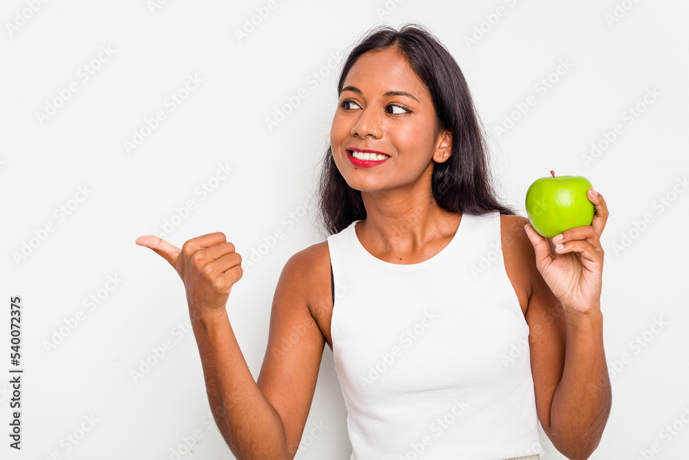 Young Indian woman holding an apple isolated on white background points with thumb finger away, laughing and carefree. Young Indian woman holding an apple isolated on white background points with thumb finger away, laughing and carefree.