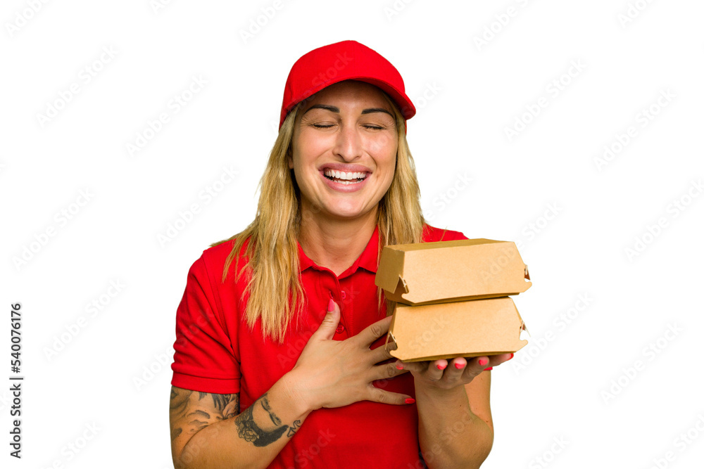 Young caucasian delivery woman holding a burger isolated on green chroma background laughs out loudly keeping hand on chest. Young caucasian delivery woman holding a burger isolated on green chroma background laughs out loudly keeping hand on chest.