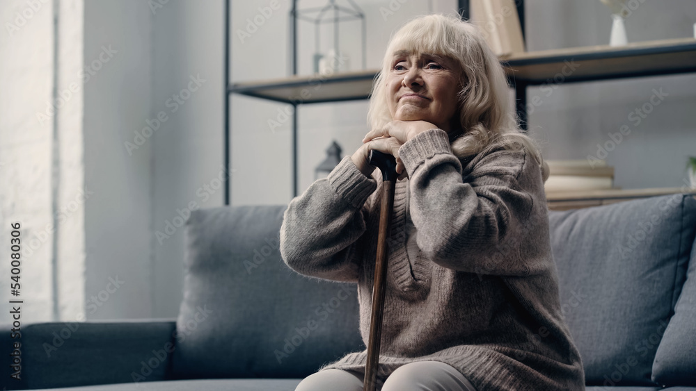 Fototapeta premium sad senior woman with dementia sitting on sofa while leaning on walking cane.