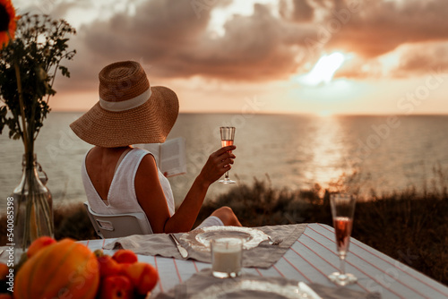 a young woman in a hat sits by the sea at sunset, reads a book and drinks champagne