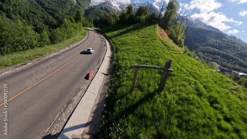 Aerial view spring sunny alpine forest resort infrastructure white sport car riding on slope serpentine road. FPV drone asphalt highway level automobile fast driving up mountain peak nature scenery 4k photo