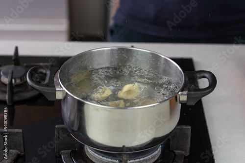 Chef cook hand making handmade tortellini ravioli with boiling water in pot saucepan on stove fire.