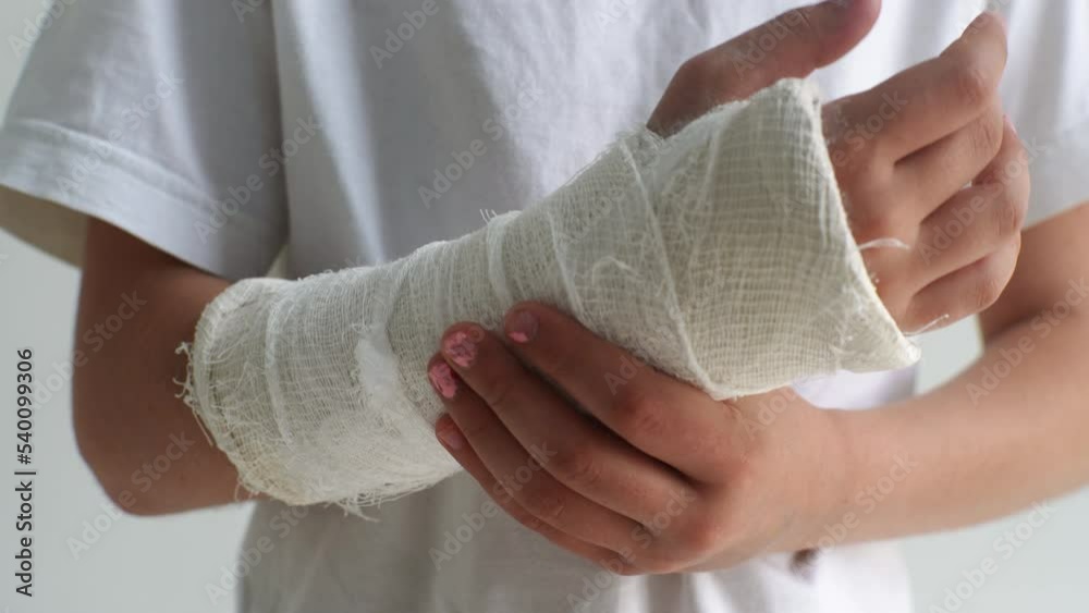 Close-up cropped shot of unrecognizable little girl with broken hand wrapped in white plaster bandage gently massaging injured forearm standing on white isolated background. Shooting in slow motion.