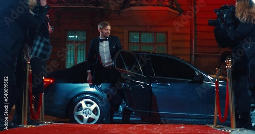 Handsome man in stylish suit helping elegant blonde woman to come out of luxury black car while arriving to event with red carpet and paparazzi around. Celebrity, fame and party concept.