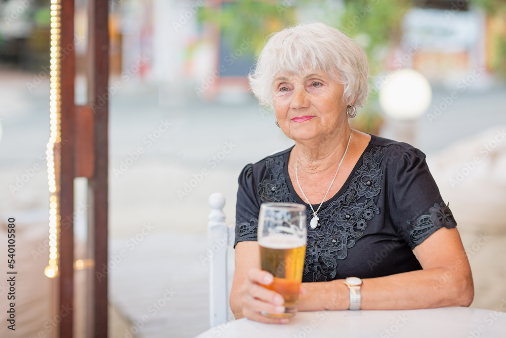 Senior woman sitting at a table in a summer cafe and drinking beer from a tall glass Senior woman sitting at a table in a summer cafe and drinking beer from a tall glass