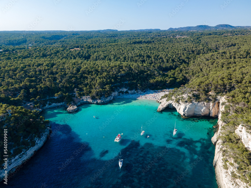 drone views of virgin beach surrounded by vegetation, with people and boats moored, enjoying a day at the beach on the coast of Menorca.
