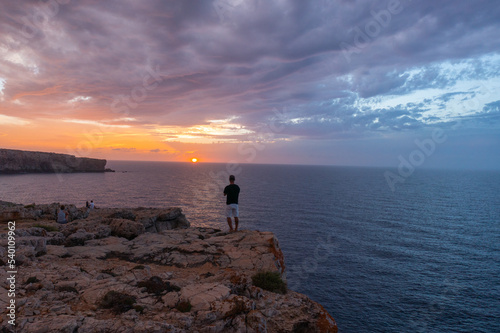 drone view with person standing on his back, watching the sunset on cliff, accompanied by more people on the coast of menorca.