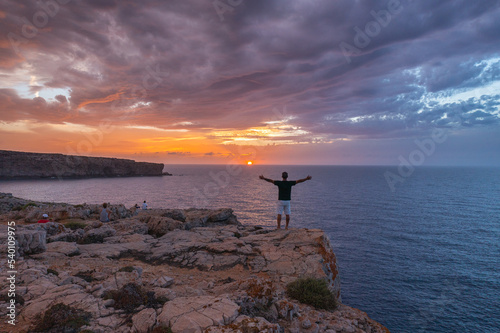 panoramic from drone, person with open arms enjoying the sunset in the company of more people on the Minorcan coast.