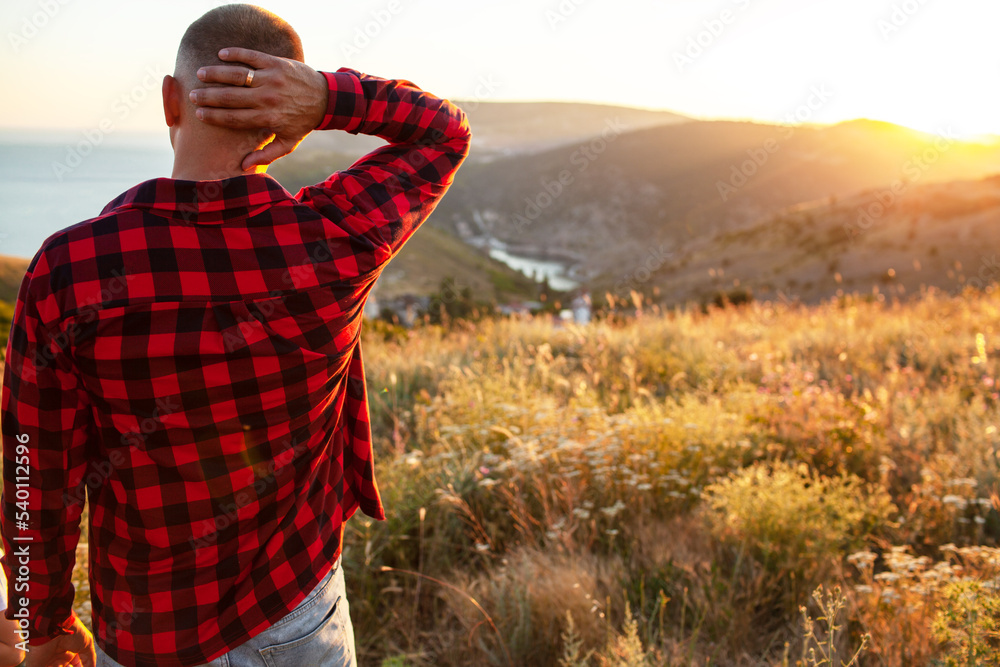 The man dressed jeans, shirt and t-shirt walkinf in the field of the grass against the  background of sunset, mountain