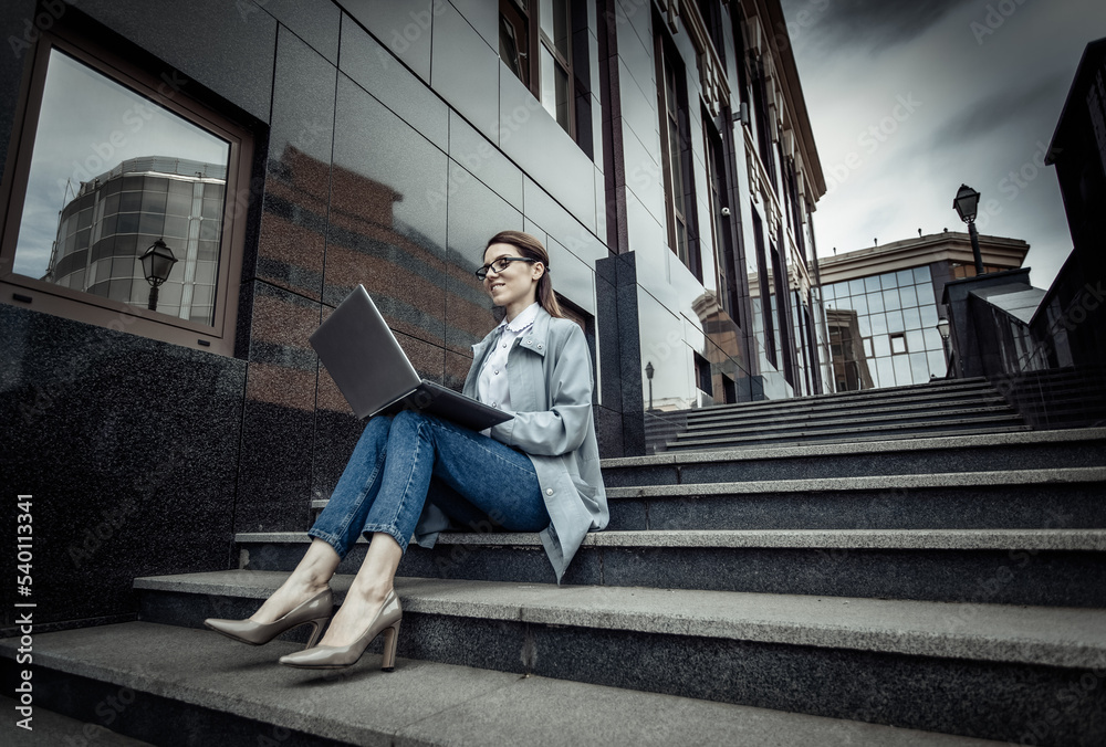 Cute Caucasian woman is using laptop while sitting on stairs outdoors. Distant work. Modern business woman. Uses gadgets. Lifestyle Cute Caucasian woman is using laptop while sitting on stairs outdoors. Distant work. Modern business woman. Uses gadgets. Lifestyle