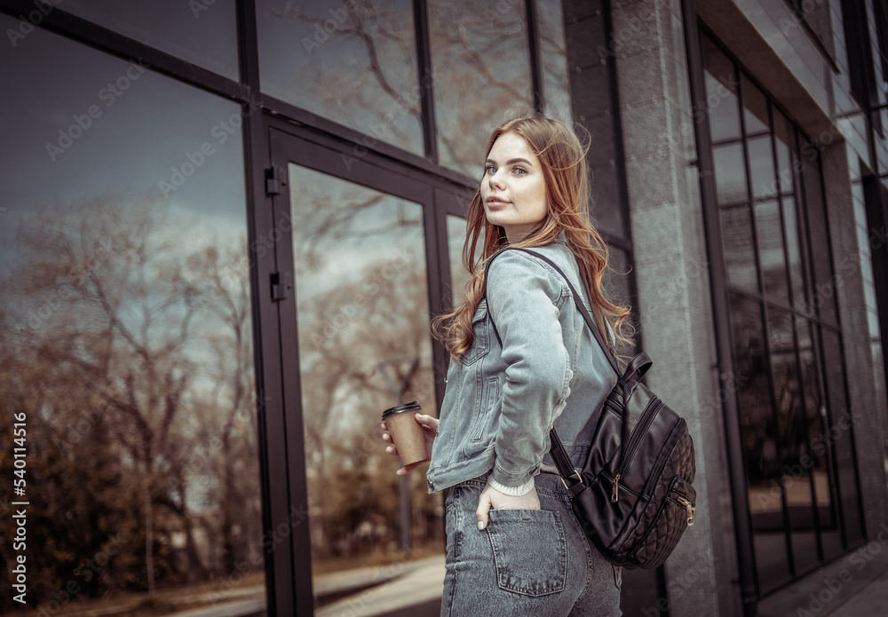 Young beautiful woman in denim jacket and holding a cup of coffee near the windows of the business center