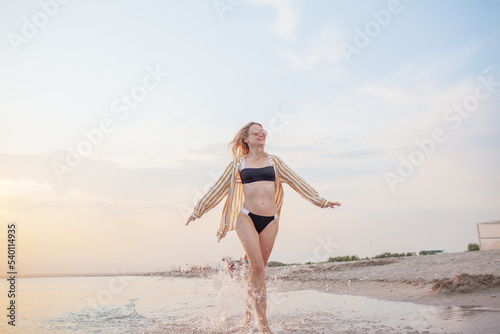 A beautiful girl is resting on the seashore against the background of sunset? the girl is wearing a swimsuit, sunglasses and a shirt