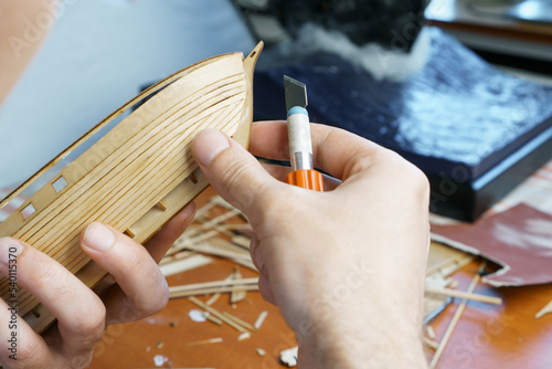Hands of man cutting out details for ship model with clerical knife from plywood. Process of building toy ship, hobby and handicraft. Table with various materials, parts and devices for work