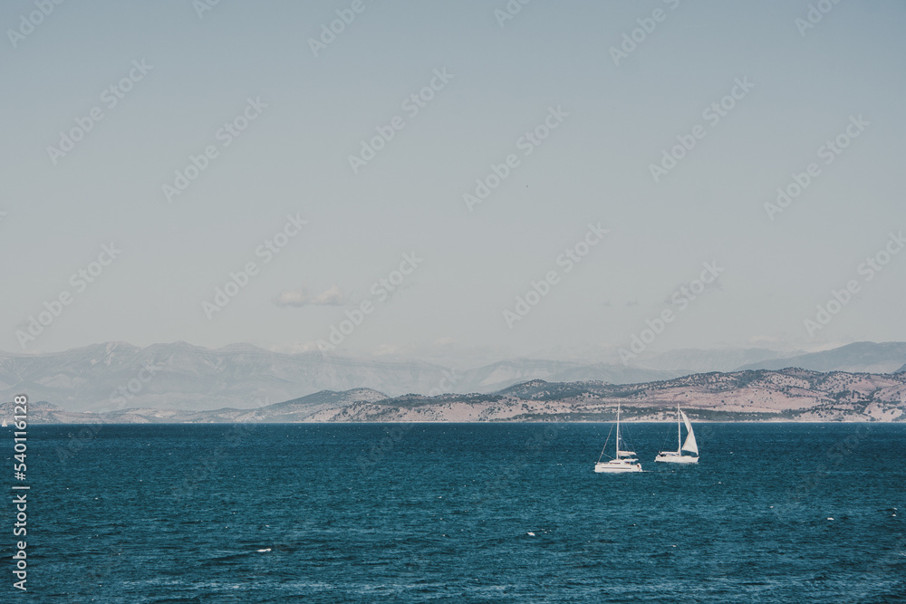 beautiful view of the Greek mountains by the blue water on which two white sailboats are sailing.