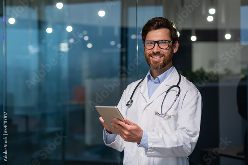 Portrait of mature and experienced doctor, man in medical coat and glasses looking at camera, doc using tablet computer, working inside modern office clinic