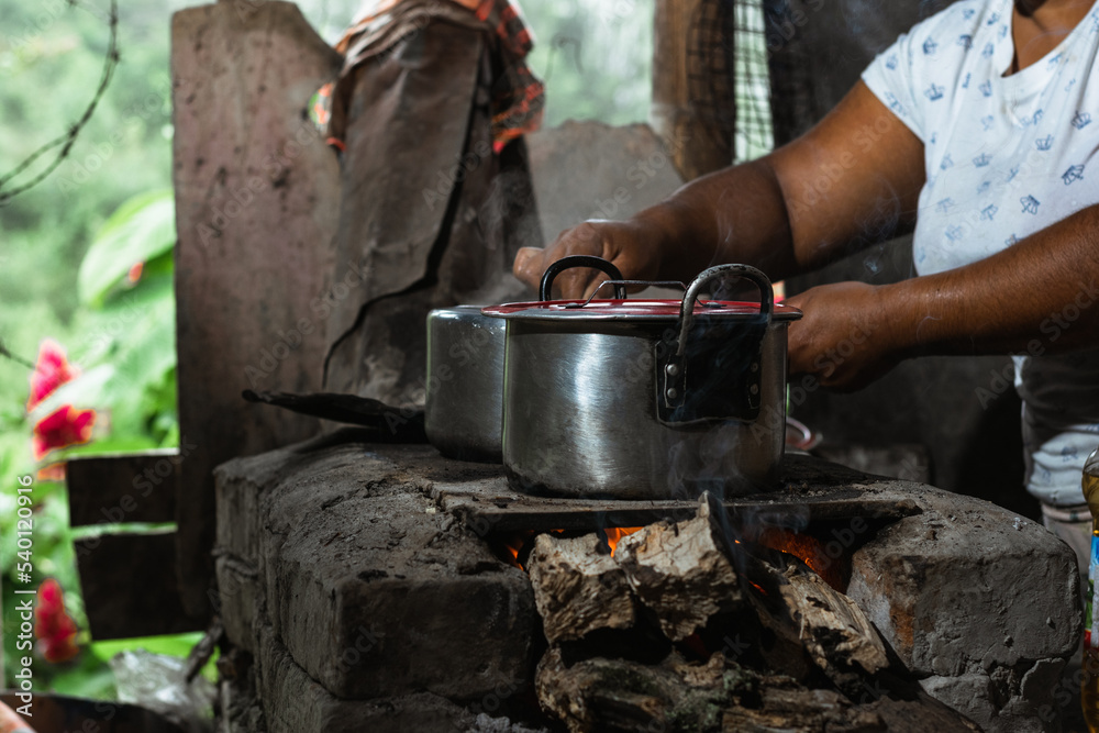 close-up of the hands of a latin woman, a brown-skinned peasant, holding an iron pot on top of a traditional stove, commonly used for cooking on a colombian farm, using wood as fuel. close-up of the hands of a latin woman, a brown-skinned peasant, holding an iron pot on top of a traditional stove, commonly used for cooking on a colombian farm, using wood as fuel.