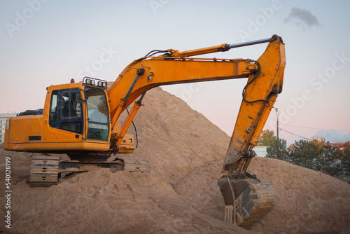 A tracked orange backhoe loader is parked on a large mountain of sand. A crawler excavator loads crushed stone and sand into a truck. Large hills and pillars of sand.