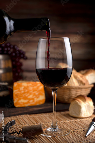 glass of red wine on wooden table with bread and cheese, bottle opener and background wooden barrel with grapes